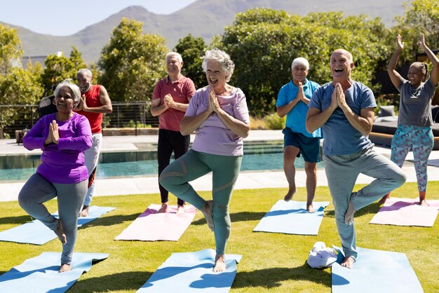 Group of active seniors practicing yoga in tree poses on mats outdoors near a pool, enjoying a sunny wellness session together.