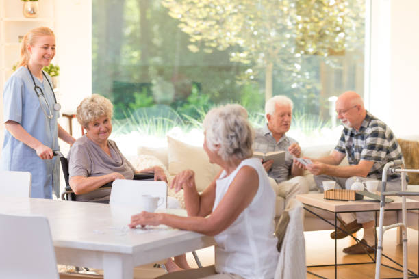 Senior residents enjoy conversation and card games in a sunny common room while a caregiver supports a woman in a wheelchair.