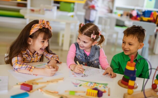Three young children smiling and drawing together at a table with crayons in a bright classroom.